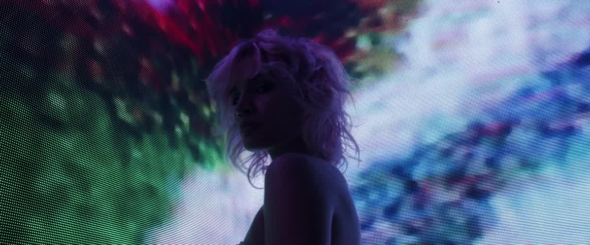 A woman in a sparkling dress standing in the corner of a video wall turns her head to gaze into the camera