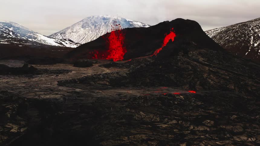 Erupting volcano spews molten hot lava into the air and oozes a river of hot lava with snow covered mountains in the distance