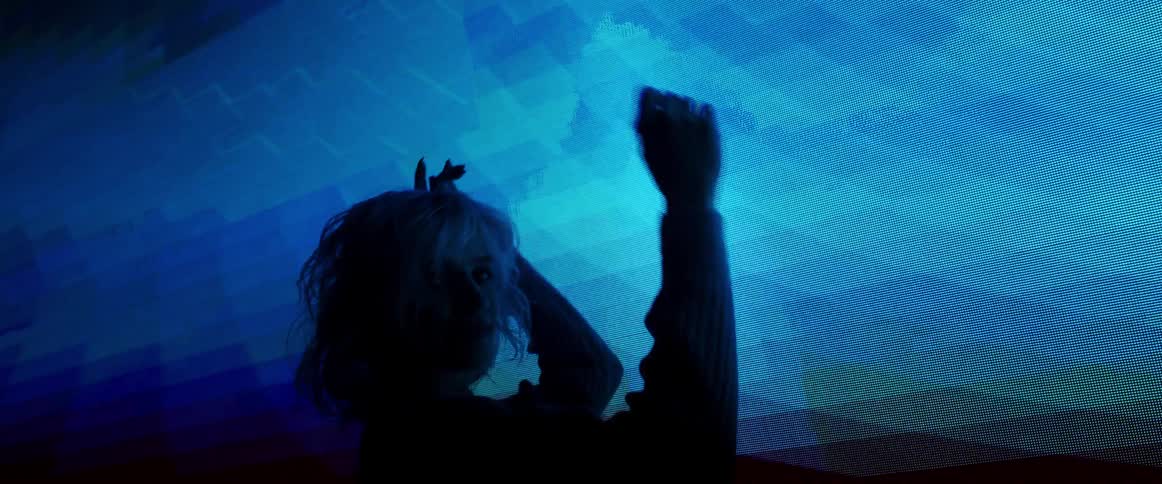 A woman with short blond hair dances in front of a large display as abstract imagery flash behind her