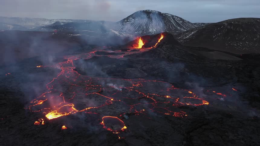 Smoke rises as an erupting volcano spews molten hot lava into the air and oozes a river of hot lava with snow covered mountains in the distance