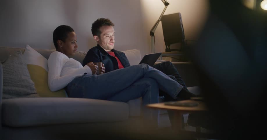 A man and woman sit together on a couch watching a laptop screen