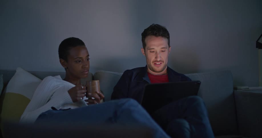 A man and woman sit in a dark room together looking at a tablet