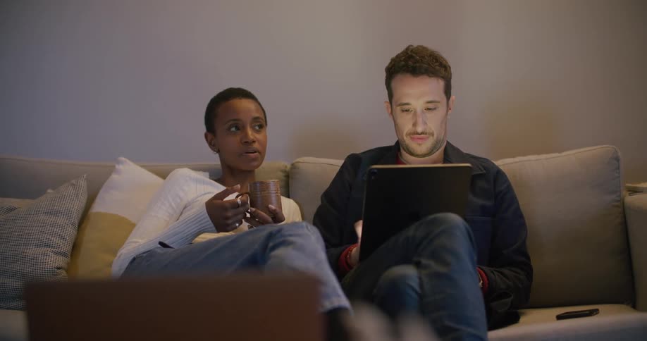 A man and a woman sitting on a couch in a living room talking to each other as the man scrolls on a tablet