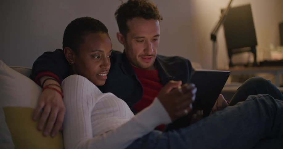 A man and a woman sitting on a couch looking at a tablet with a wall in the background