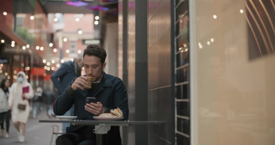 Man drinks coffee while checking his smartphone sitting in a table on the city streets