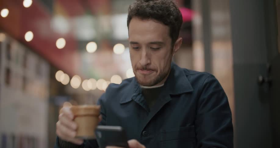A man sits at a table with a sandwich and takes a drink of his beverage while looking at his phone