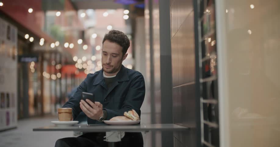 A man sits outside a restaurant and takes a drink of his beverage as he looks at his cell phone
