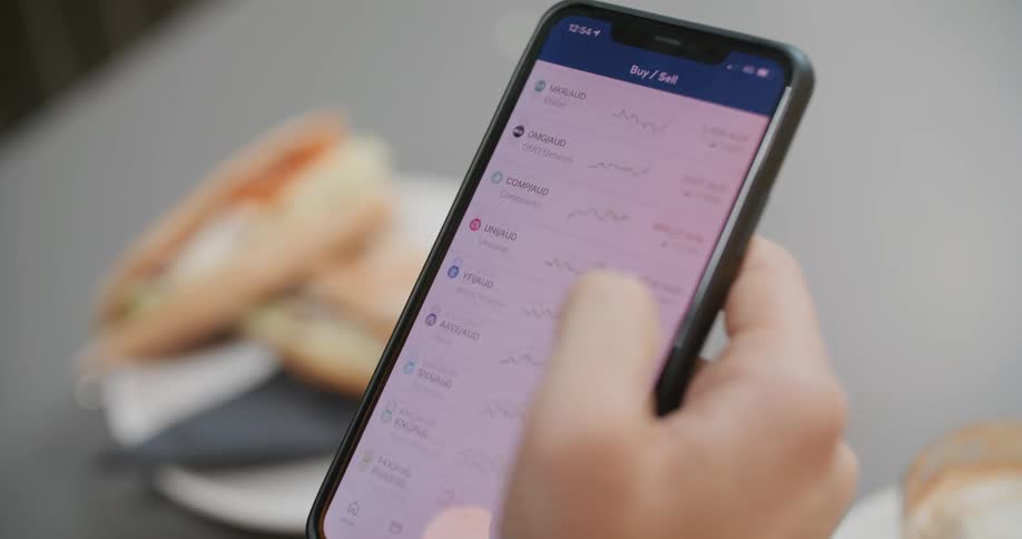 A person uses their thumb to scroll a list of stocks on a smartphone while sitting at a table