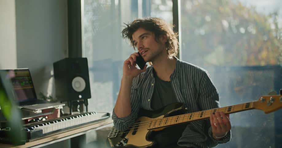 A guitar player hangs up a phone call and resumes recording music on his laptop