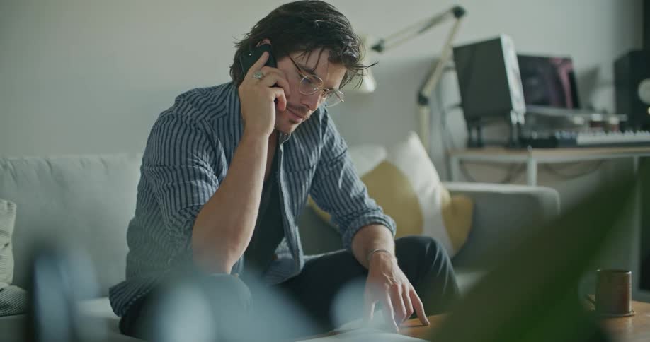 A man sitting on his couch with laptop open on the coffee table answers and talks on his phone