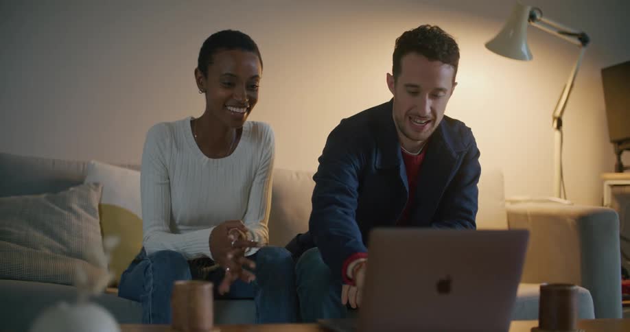 A man and woman smile and talk as they sit on a couch with a laptop in front of them