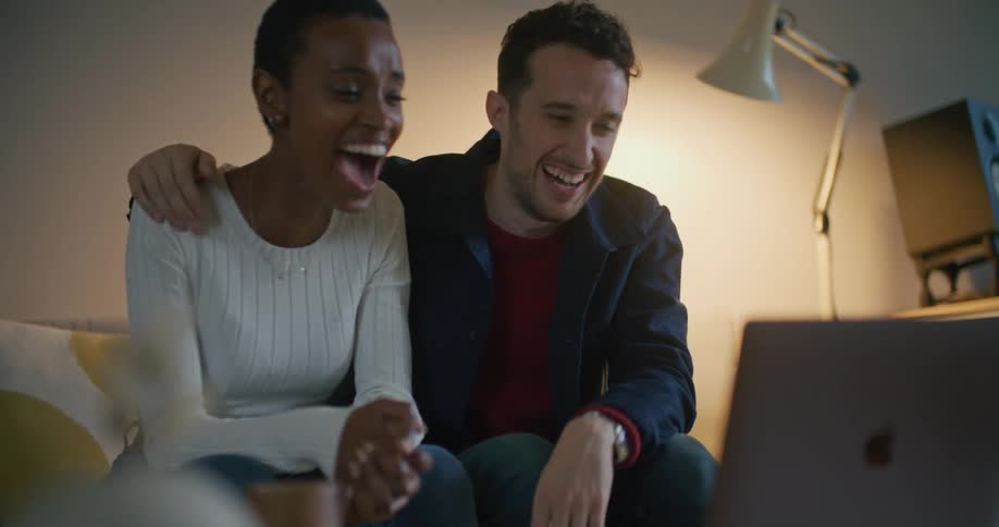 A woman and man laugh as they watch the screen of a laptop while sitting together on a sofa in a house