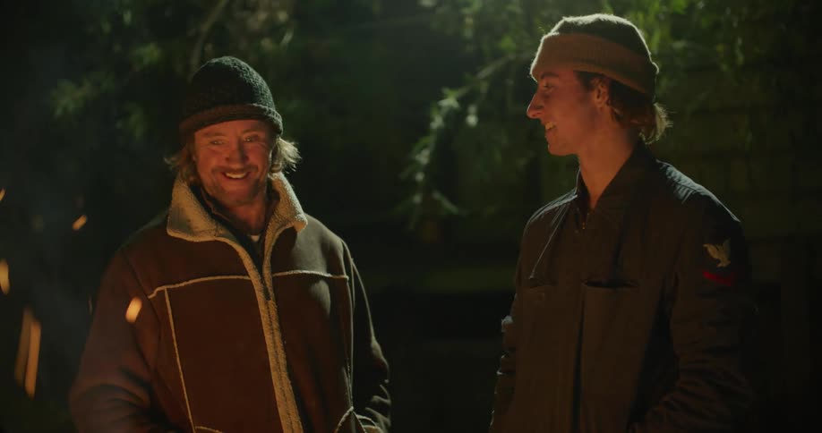 Two men wearing coats and knit hats talk and laugh while standing outdoors at night next to fire shooting out sparks