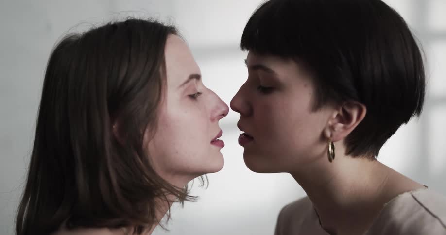 Two teenage girls sit facing one another and remove their shirts and then lean in for a kiss and hug one another