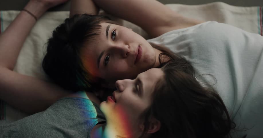 A pair of young women speak to one another while resting next to one another on a bed during the day