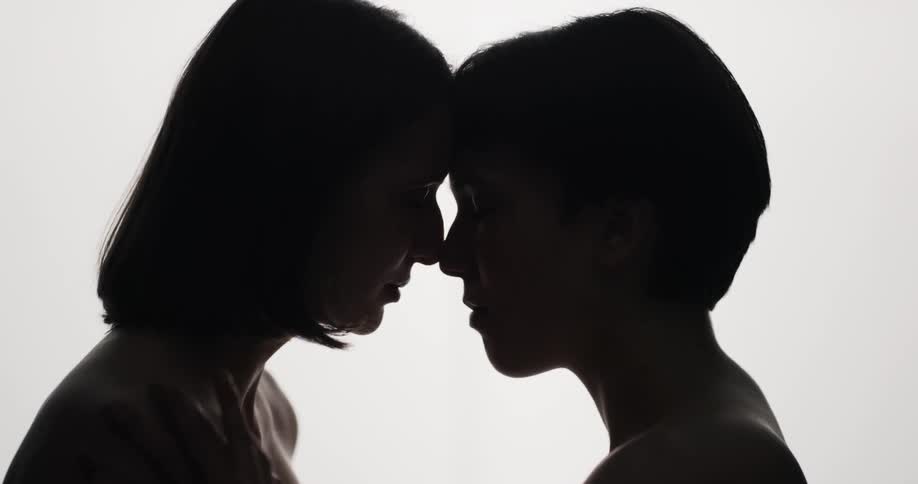 Two teenage girls stand naked with their heads together as one rubs the others shoulder