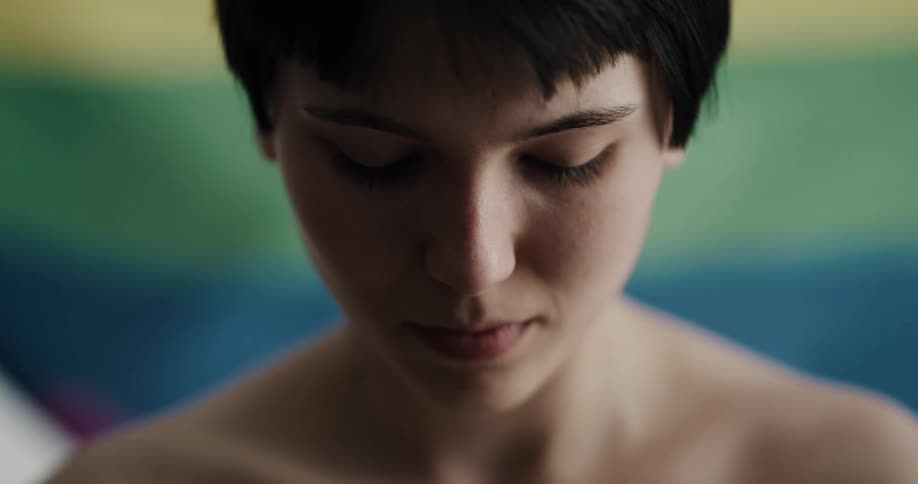 A young shirtless woman stands in front of a waving lgbt pride flag with her head down