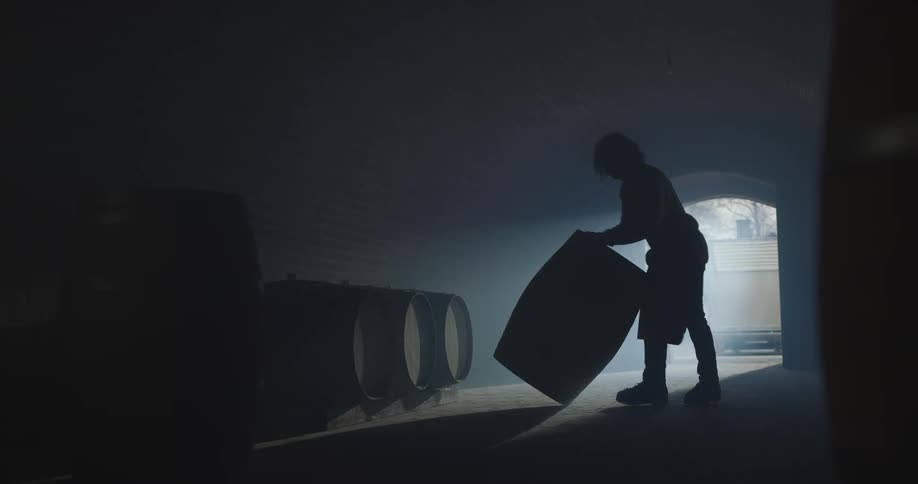 A man rolls a barrel into a dark warehouse and stores it there before leaving and closing the door
