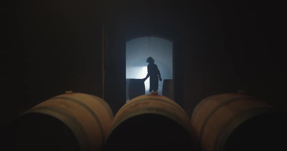 A man carefully rolls a large wooden barrel through a dark cellar before placing it on its end