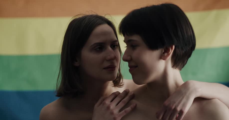 Two women in front of a pride flag as one woman embraces the other woman from behind and they smile at each other
