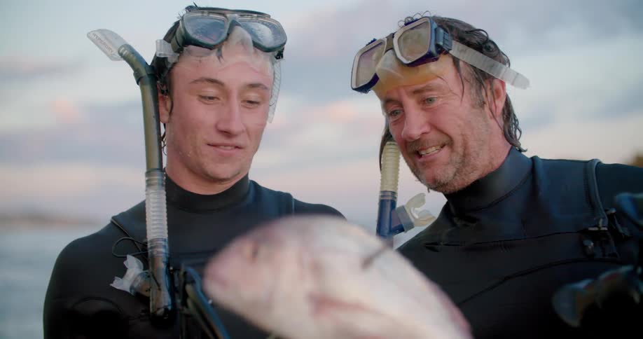 Two men with scuba gear looking at a fish with the sky and clouds in the background