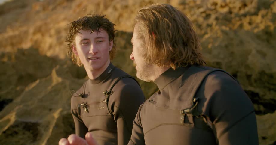 Two men talking while sitting on a sandy beach in wet suits