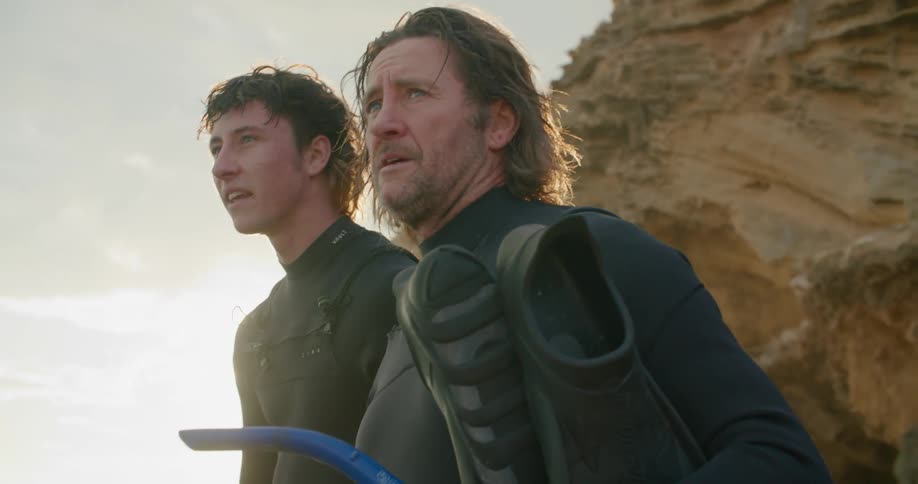 A father and son wearing surf gear standing outside in front of a flare of sunlight next to a high rocky cliff