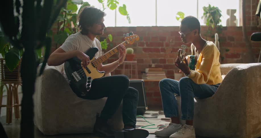 A man and a woman in a room sitting and playing guitars with a wall and window in the background