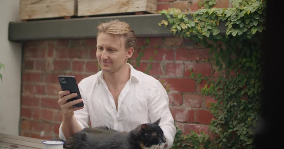 A man sets his mug down and pets his cat while using his smartphone and sitting outdoors