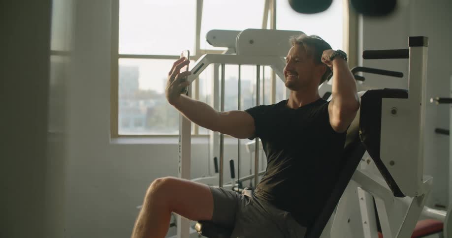 A guy smiles while taking a selfie and flexing his muscles while sitting on gym equipment