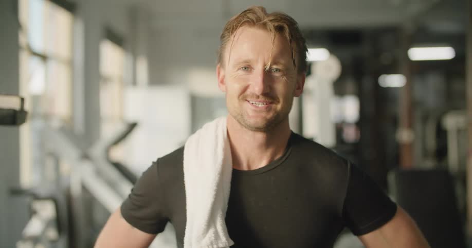 A sweaty man smiles as he poses in a gym with a towel draped over his shoulder