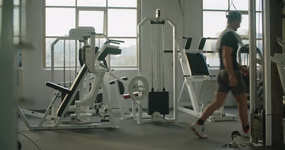 A man walking in a gym and throwing a towel over his shoulder with equipment and windows in the background