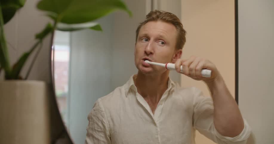 A man brushes his teeth using an electric toothbrush and then he spits and walks away
