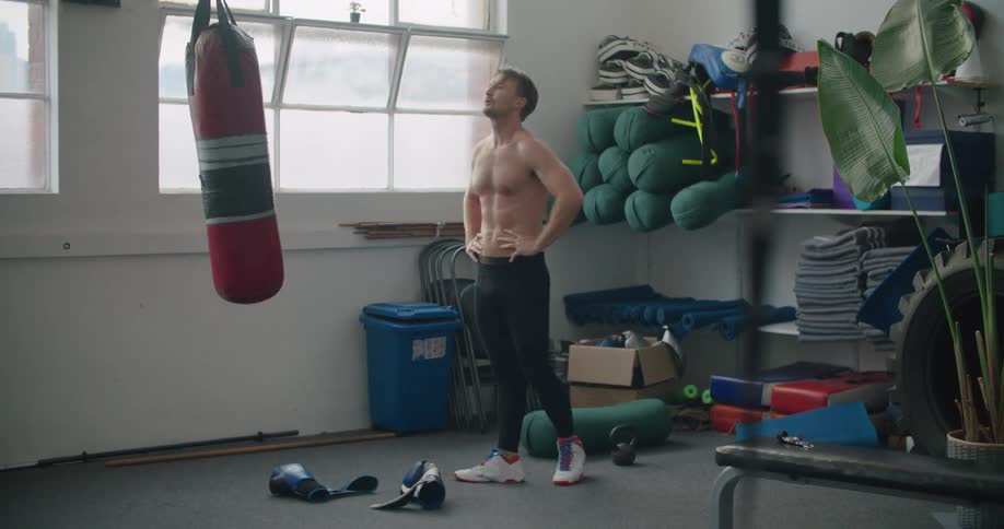 A shirtless man takes off his boxing gloves after exercising using a punching bag