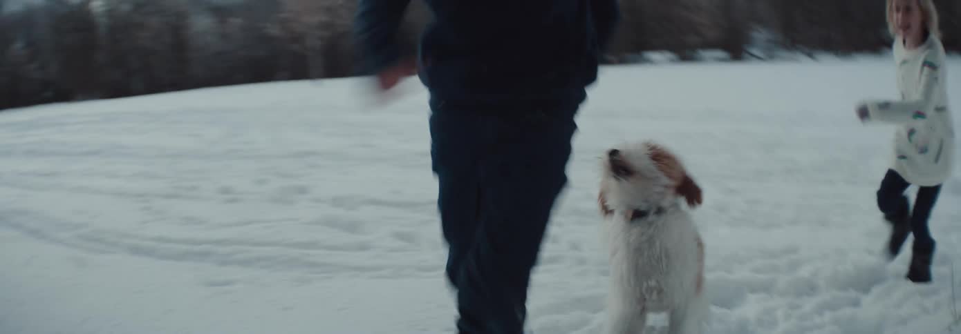 Boy and girl run around a tree with a white brown dog in the snowy field with snow capped mountains