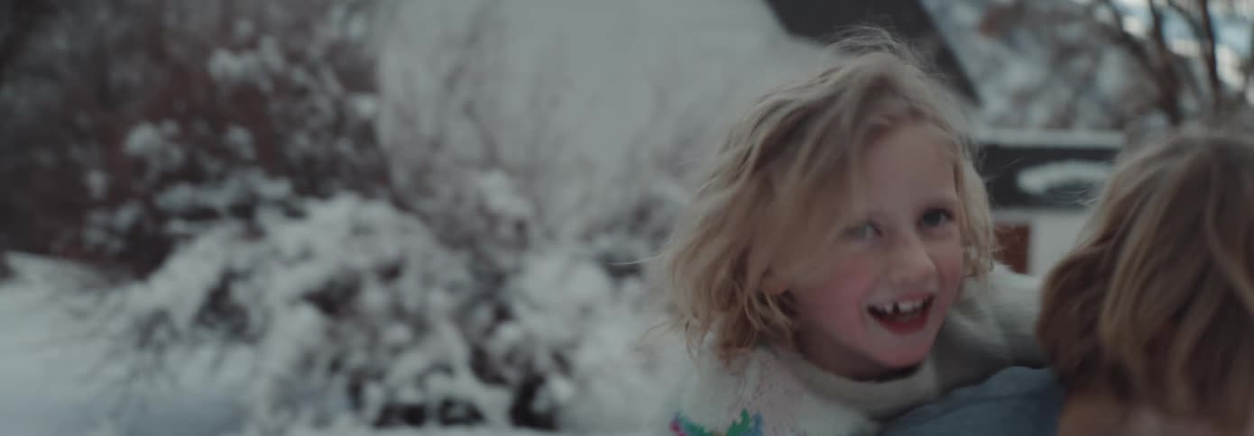 A boy standing in snow with a girl on his back with a house trees the sky and clouds in the background