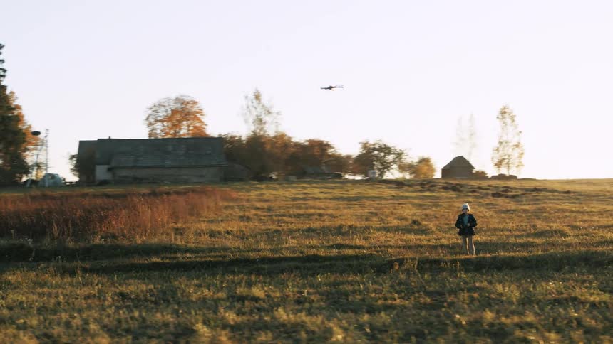 Child wearing a knit hat and a jacket is watching a drone as it flies over a field