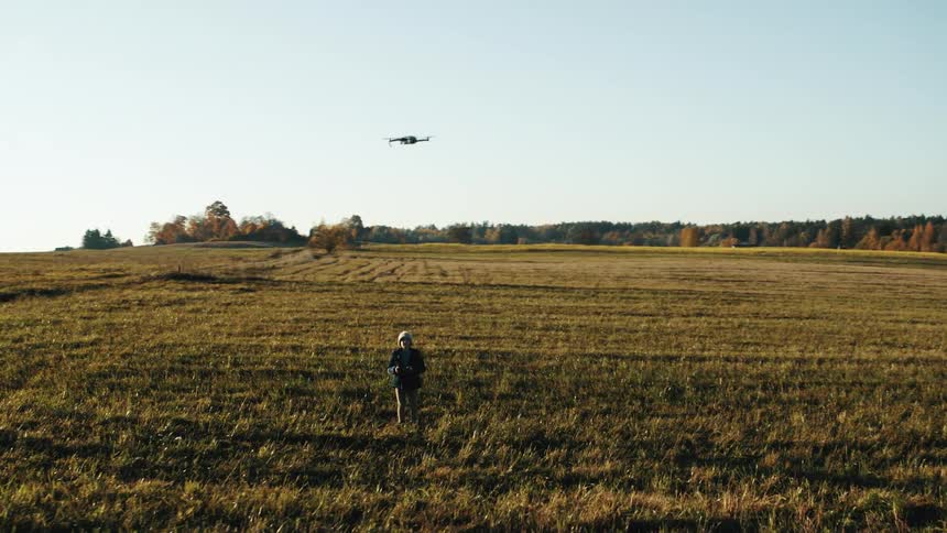 Child wearing a knit hat and a jacket is flying a drone in an open field with fall colored trees in the distance