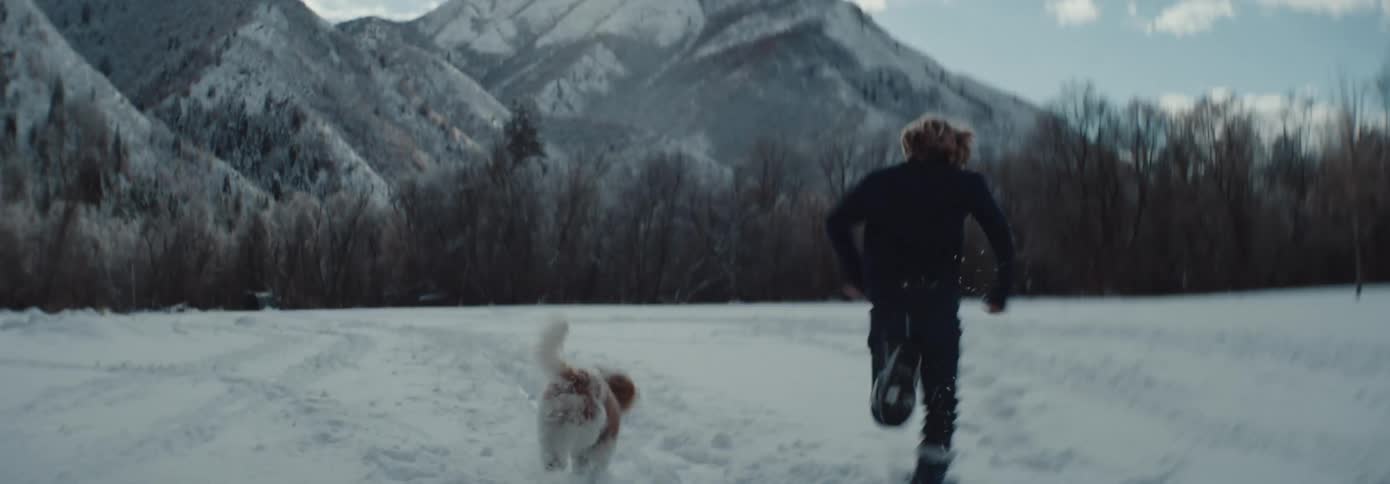 Brother and sister run trough snow near farmhouse toward leafless forest and mountain with pet dog