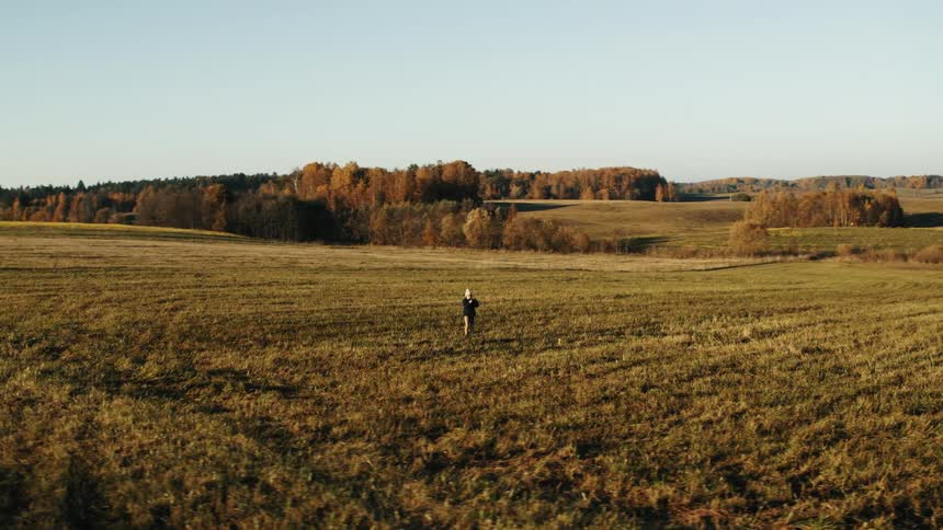 Child wearing a knit hat and a jacket is standing an open field with fall colored trees in the distance