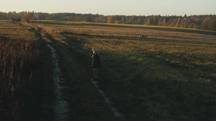 A person with a hat and jacket flying a drone on a grassy field with the sky and clouds in the background