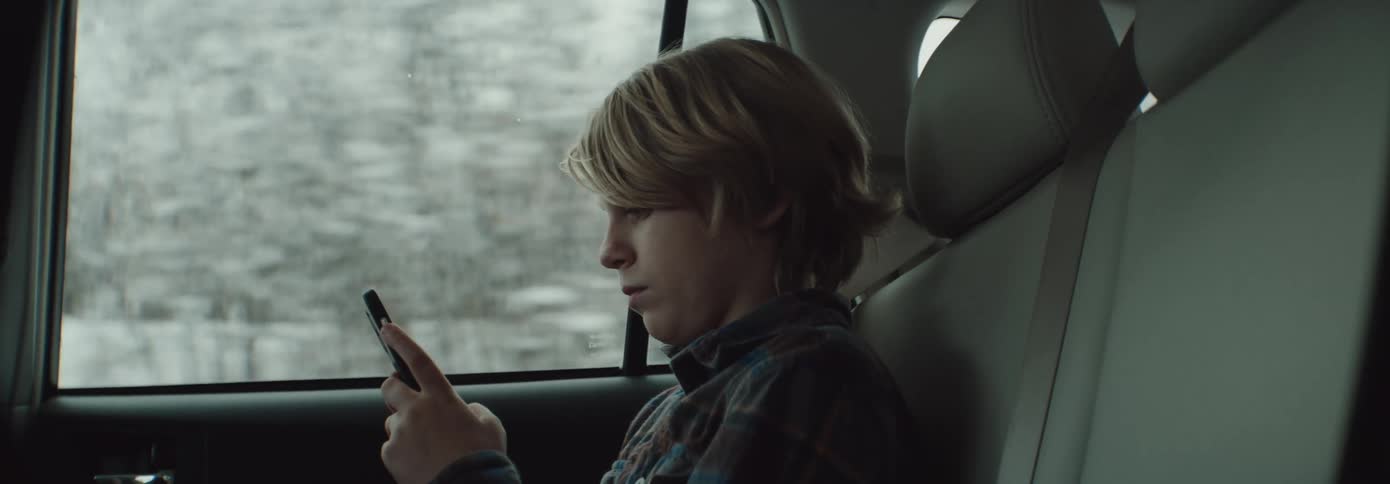 A young boy sits in a car and texts on a cell phone