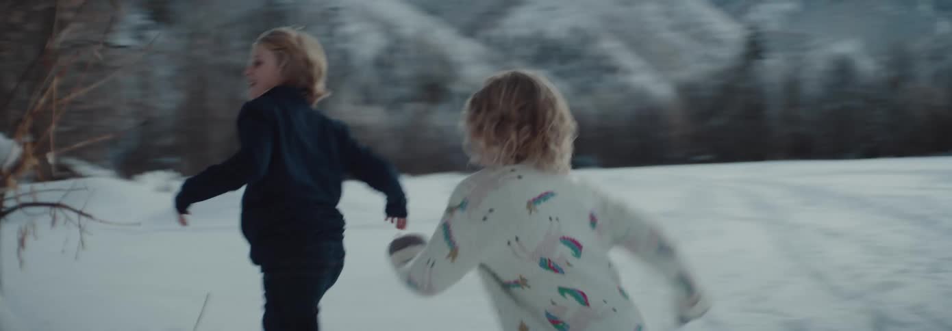 Boy and girl run around a tree next to a building in the snowy field