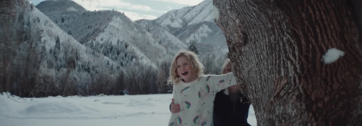 A boy and a girl dodging behind a tree from a snowball with trees the mountain range the sky and clouds in the background