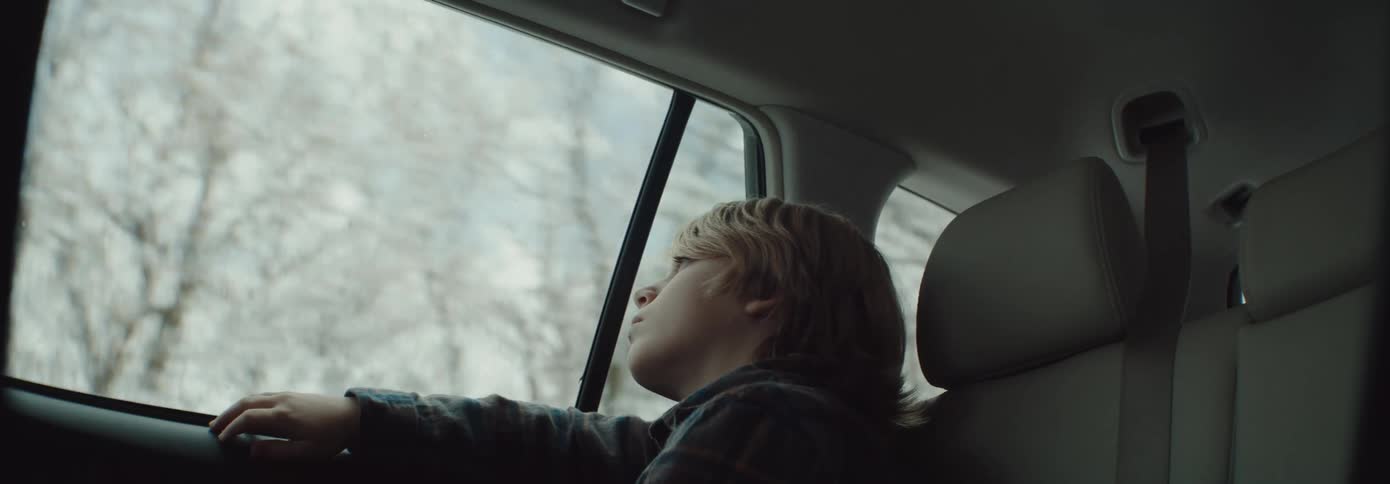 A boy sitting in a vehicle looking out the window with trees a mountain range the sky and clouds in the background