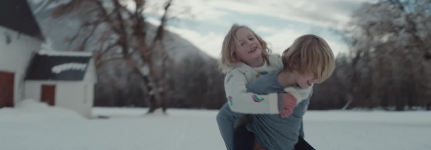 A boy playfully spins around with his younger sister on his back in a snowy field during the day