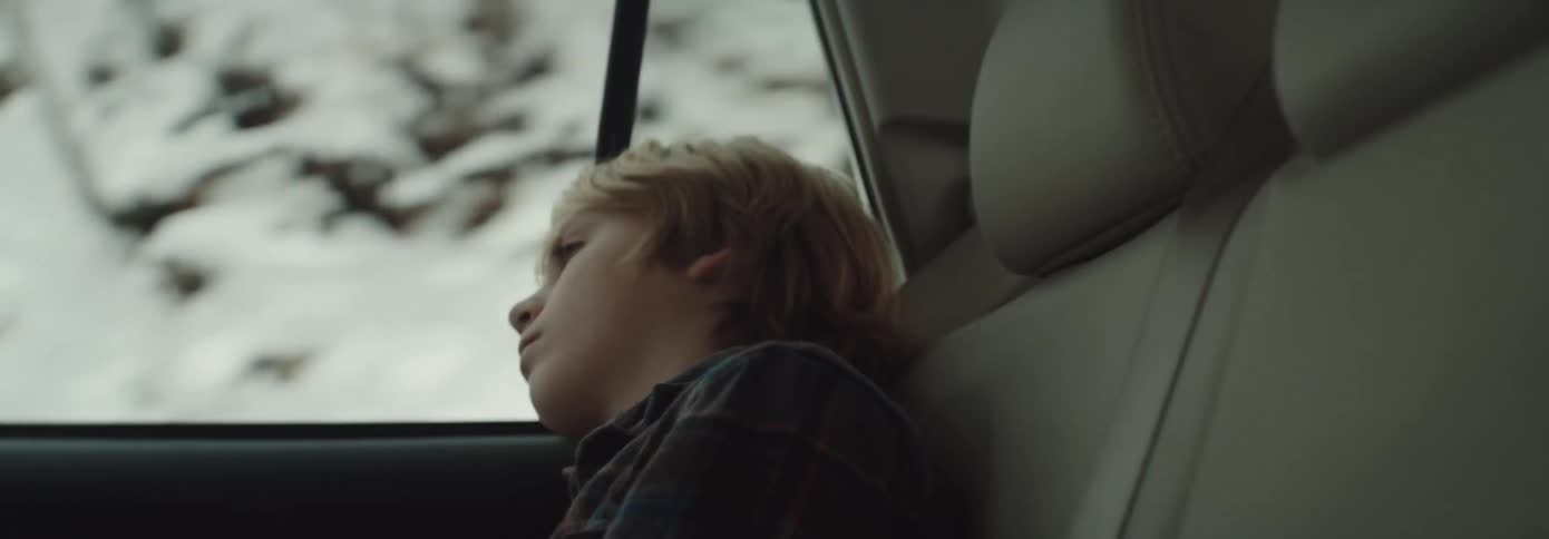 Side view of blonde boy looking out window of moving car through snowy forest