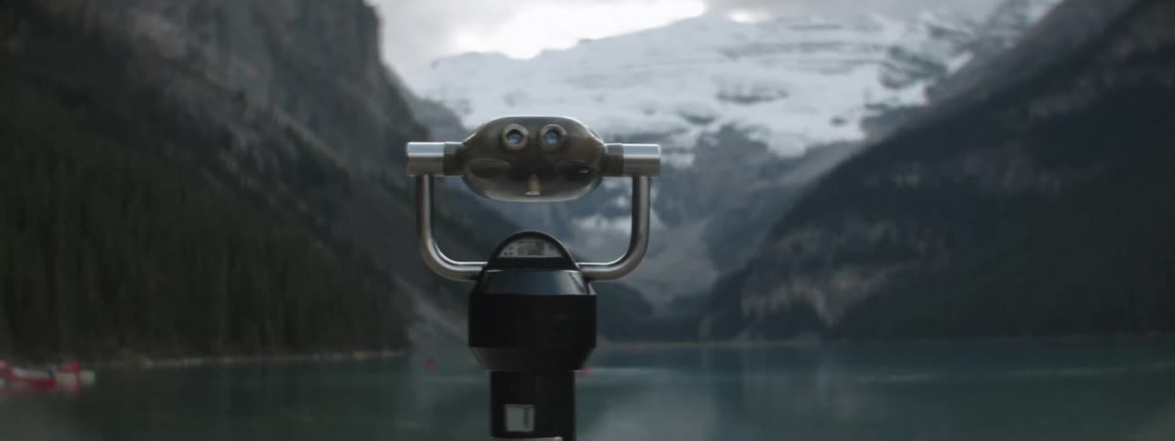 A telescope on the shore with people in boats on the water and mountains in the distance