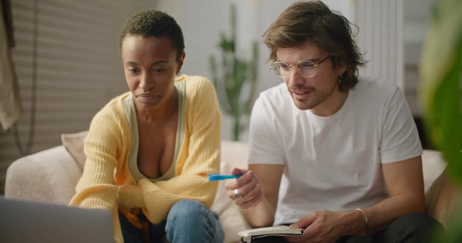 A man and a woman sitting in a room looking at a laptop with a wall and a plant in the background