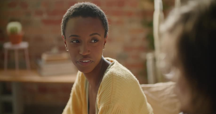 A woman sits indoors on a couch while she speaks with a male companion